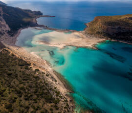 A wide panoramic view of Balos Lagoon, revealing the gentle contrast between sea, sand, and rugged coastline. Panoramic aerial view of Balos Lagoon in Crete with layered coastline, turquoise water, and distant horizon.