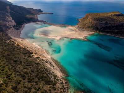 A wide panoramic view of Balos Lagoon, revealing the gentle contrast between sea, sand, and rugged coastline. Panoramic aerial view of Balos Lagoon in Crete with layered coastline, turquoise water, and distant horizon.