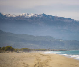 Where turquoise water meets snow-covered mountains – a rare winter moment on the island of Crete. Winter landscape in Crete with beach, turquoise water and snow-capped mountains in the background.